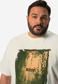 Camiseta de algodón blanca con un diseño gráfico en verde y naranja que incluye la palabra "UNTAMED" y elementos botánicos. Cuello redondo y mangas cortas.