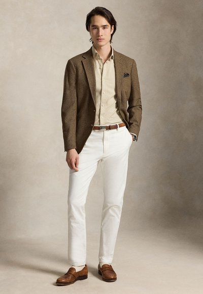 Jeune homme debout, une main dans la poche, portant un blazer marron, une chemise beige à rayures, un pantalon blanc, une ceinture marron et des mocassins marron.