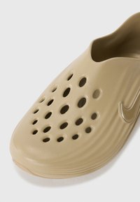 Gaišs beidžu krāsas slip-on apavs ar vairākiem apļveida ventilācijas caurumiem, gludu plastmasas tekstūru un viegli pamanāmu Nike logotipu sānos.