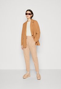 Beige Blazer über einem cremefarbenen Oberteil, kombiniert mit hochgeschnittenen beigen Hosen und cremefarbenen Plateausandalen. Dunkle Sonnenbrillen vervollständigen das Outfit.