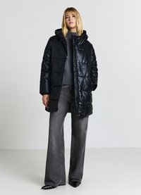 Schwarzer wattierter Steppmantel mit Kapuze, mit glatter Textur und gestepptem Muster. Getragen über einem grauen Strickpullover und grauen weit geschnittenen Hosen.