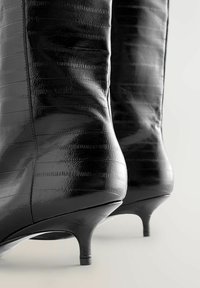 Stivaletti neri in pelle con finitura lucida, caratterizzati da un motivo testurizzato, punte appuntite e tacchi a stiletto di 5 cm.