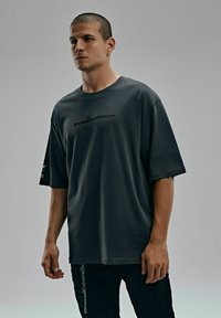 Ciemnoszara, oversize t-shirt z krótkim rękawem, z logo na przodzie i wyraźnym czarnym tekstem na rękawie. Wykonana z miękkiego materiału bawełnianego.