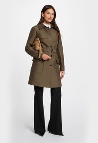 Olijfgroene mantel met riem, een kraaghalslijn, knoopsluitingen en voorzakken, gecombineerd met zwarte wijde broek en een bruine franjes tas.