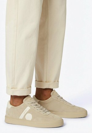 Beige suède lage sneakers met witte cirkel- en streepaccenten, gedragen met opgerolde crèmekleurige broek.