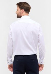 Weißes Langarmhemd aus glattem Stoff mit klassischem Kragen und dunklen Knopfakzenten an den Manschetten, getragen mit dunklen Hosen.