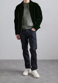 Grön sammetsjacka över en grå tröja, kombinerad med mörka jeans och vita sneakers. Jackan har dragkedja och uppvikt krage.