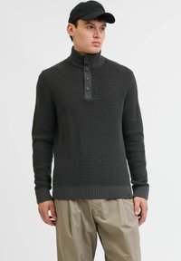 Suéter de punto gris oscuro con cuello alto, tapeta de cuatro botones y puños acanalados. Combinado con pantalones sueltos de color beige claro.