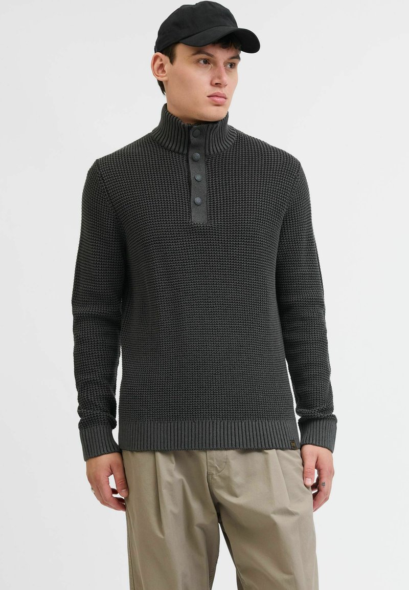 Suéter de punto gris oscuro con cuello alto, tapeta de cuatro botones y puños acanalados. Combinado con pantalones sueltos de color beige claro.