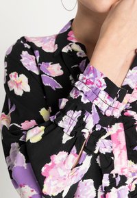 Blumenbluse mit schwarzem Hintergrund, die rosa und lila Muster zeigt. Bedeutende Details sind die gerafften Ärmel und die geknöpfte Manschette.