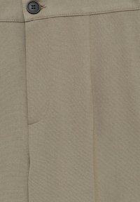 Gros plan sur un pantalon en tissu beige avec un seul bouton noir, des coutures visibles et un contour de poche partiellement cousu.
