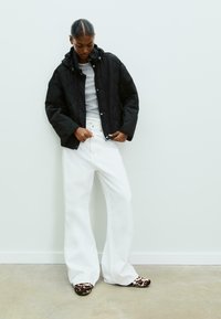 Veste matelassée noire avec capuche, top gris dessous, pantalon large blanc et chaussures brunes à motifs. Design simple avec peu de matériel.
