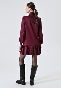 Burgunderfarbenes langärmliges Kleid mit Rüschensaum, aus glattem Stoff, kombiniert mit schwarzen knielangen Stiefeln mit goldenen Hardware-Details.