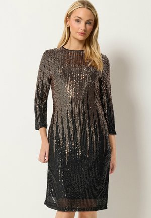 Femme aux longs cheveux blonds portant une robe noire et dorée à sequins ajustée jusqu'aux genoux avec des manches trois-quarts, debout contre un mur clair uni.