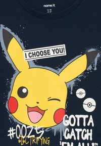 Gul Pikachu-ansikte med röda kinder som blinkar, texten "JAG VÄLJER DIG!", två Poké Bollar, och fraserna "#0025 elektrifierande" och "MÅSTE FÅNGAS ALLA" på marinblått tyg.