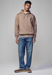 Hnedá mikina s kapucňou a predným vreckom, nosená cez biele tričko; v kombinácii s modrými džínami a hnedými slip-on topánkami so šnúrkami.