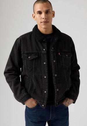 Black denim jacket with a fuzzy collar, featuring button closures, chest pockets, and a relaxed fit. Paired with blue jeans.