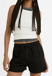 Bílý žebrovaný crop top s šedým výstřihem, v kombinaci s černými kraťasy na stažení. Oba kusy mají ležérní, uvolněný střih.