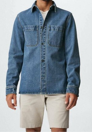 Person, die ein blaues Denimhemd mit zwei Brusttaschen und beige Shorts trägt, steht vor hellem Hintergrund.