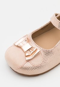 Scarpa per bambini di colore rosa chiaro con un design perforato, dotata di un'accento a fiocco in oro metallico e una punta in suede morbido. Forma arrotondata.