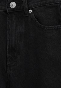 Gros plan sur la poche d'un jean noir avec un bouton argenté et des rivets sur un tissu texturé.