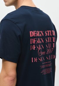 Σατινένιο μπλουζάκι από βαμβάκι σε ναυτικό χρώμα με ροζ σχέδιο κειμένου στην πλάτη, συμπεριλαμβανομένων των "DESIGN STUDIOS" και "Από το 2007," με χαλαρή γραμμή και κοντά μανίκια.