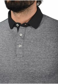 Šedé polo tričko s černým límcem, se třemi stříbrnými knoflíky, texturovaná látka a jemný vzor po celém povrchu.