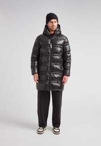 Hombre con gorro negro, abrigo de plumas largo y brillante negro con capucha, pantalones negros y zapatillas en blanco y negro, de pie contra un fondo liso.