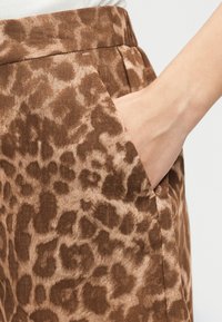 Main à l'intérieur de la poche latérale d'un pantalon marron imprimé léopard avec une ceinture froncée, sur un fond neutre.