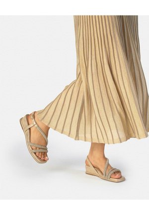 Piernas femeninas con falda plisada beige y sandalias de cuña doradas con múltiples tiras delgadas, caminando sobre un fondo liso.