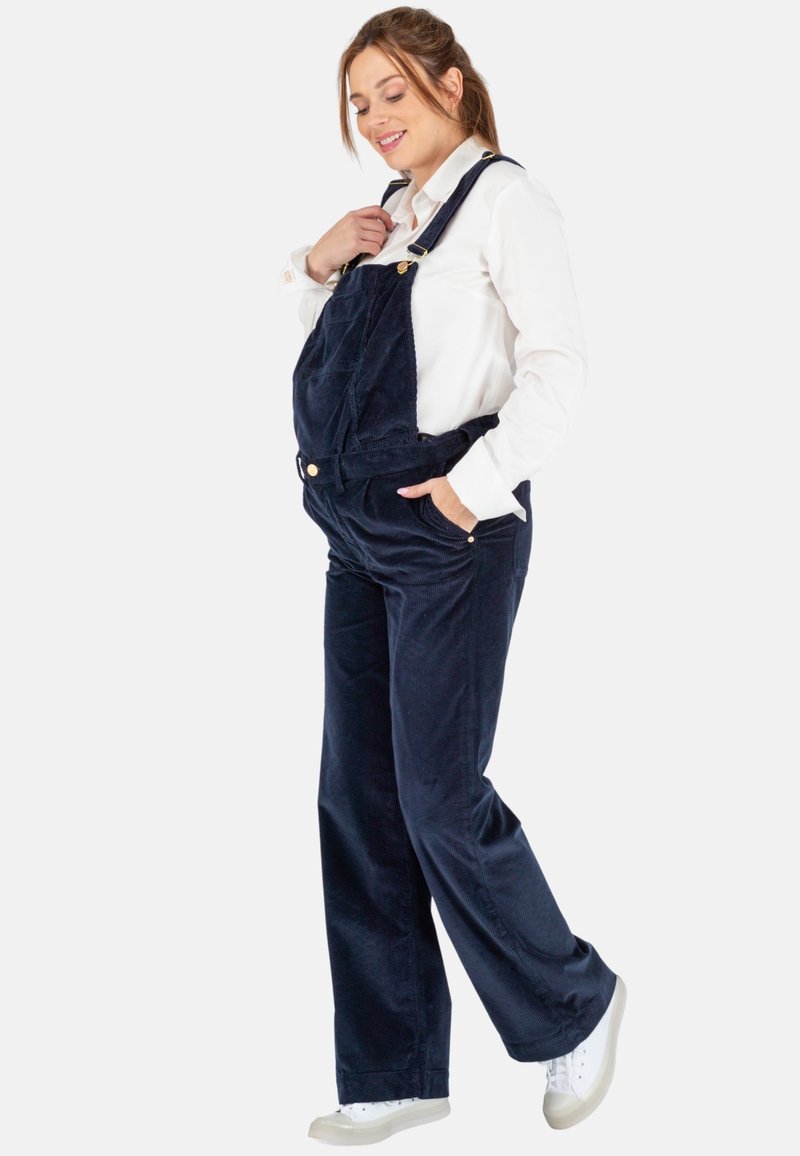 Navy corduroy overall met een relaxte pasvorm, verstelbare banden, voorzakken en details van messing, gecombineerd met een witte blouse en sneakers.