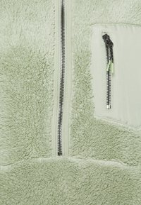 Tessuto in pile verde chiaro con due chiusure a zip nere e verdi, una verticale sulla tasca sul petto e una al centro davanti.