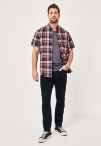 Short-sleeve plaid shirt in red, blue, and white, with a button-up design. Worn over a gray t-shirt, paired with dark jeans and black sneakers.