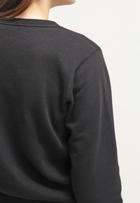 Svart bomullströja med rund halsringning, synlig sömnad och en mjuk yta, visad bakifrån. Enkel och minimalistisk design.