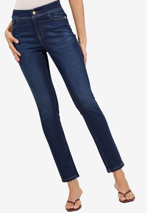 Žena nosící tmavě modré slim-fit džíny a hnědé sandály s otevřenou špičkou, stojící s jednou nohou mírně pokrčenou.