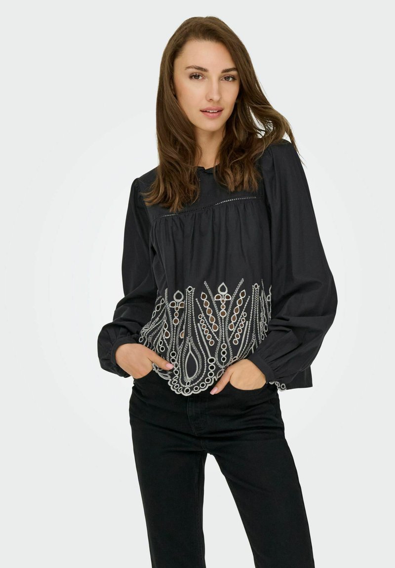 Blouse noire à manches bouffantes longues, présentant une broderie blanche complexe et des découpes circulaires à l'ourlet, fabriquée à partir d'un tissu doux.