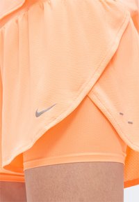 Αθλητικό σορτς σε ροδακινί χρώμα με ενσωματωμένη συμπιεστική στρώση, με μικρό γκρι λογότυπο Nike κοντά στο κάτω μέρος στο μηρό ενός ατόμου.