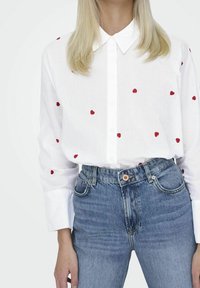 Chemise blanche à boutons avec broderie de cœur rouge, assortie à un jean en denim bleu taille haute. Tissu en coton avec une texture lisse.