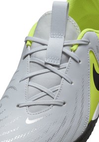 Zapatilla deportiva de color gris claro con una textura suave, que presenta detalles en negro, elementos reflectantes y detalles en amarillo neón en la lengua y los cordones.