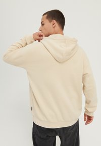 Sudadera beige con capucha, hecha de tela suave, con un ajuste relajado, puños acanalados y capucha con cordón. Etiqueta negra visible en el dobladillo.