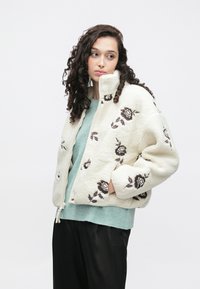 Krema flis jakna s smeđim cvjetnim vezom, visokim ovratnikom i zatvaračem na patent. Uparen s mint zelenim džemperom i crnim hlačama.