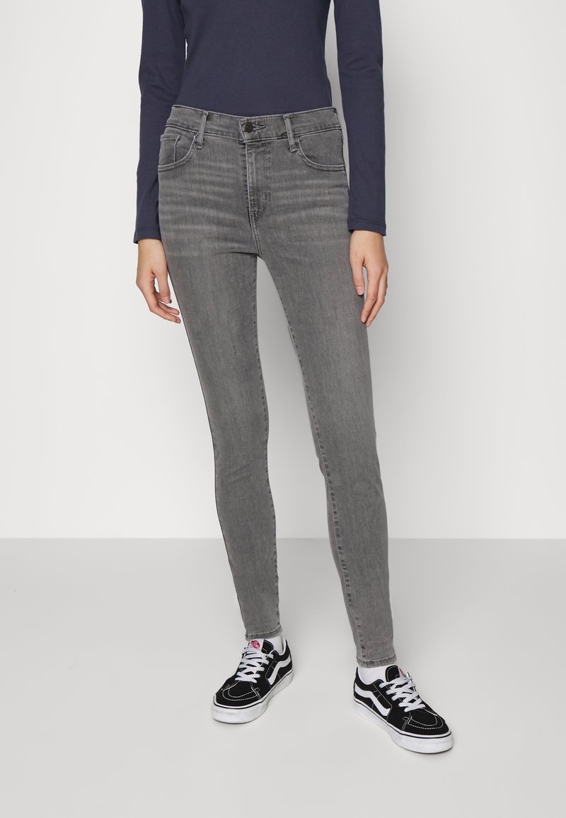 Femme portant un jean gris ajusté, des baskets noires et blanches, et un haut bleu marine à manches longues, debout devant un fond blanc uni.