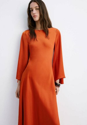 Mujer con cabello largo y castaño que lleva un vestido de satén naranja de manga larga, de pie frente a una pared blanca lisa.