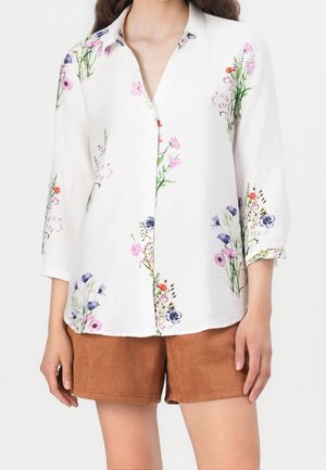 Biała koszula na guziki z lekkiego materiału, ozdobiona kolorowymi kwiatowymi wzorami w odcieniach różu, niebieskiego i zieleni; zestawiona z brązowymi szortami.
