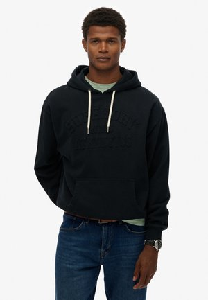 Zwarte hoodie met reliëf logo "Superdry", trekkoord-hals, voorzak en zachte textuur, gecombineerd met blauwe jeans en een horloge.
