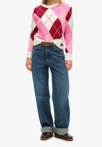 Persona che indossa un maglione argyle rosa, rosso e bianco, jeans blu arrotolati con una cintura marrone e scarpe marroni, in piedi davanti a uno sfondo bianco.