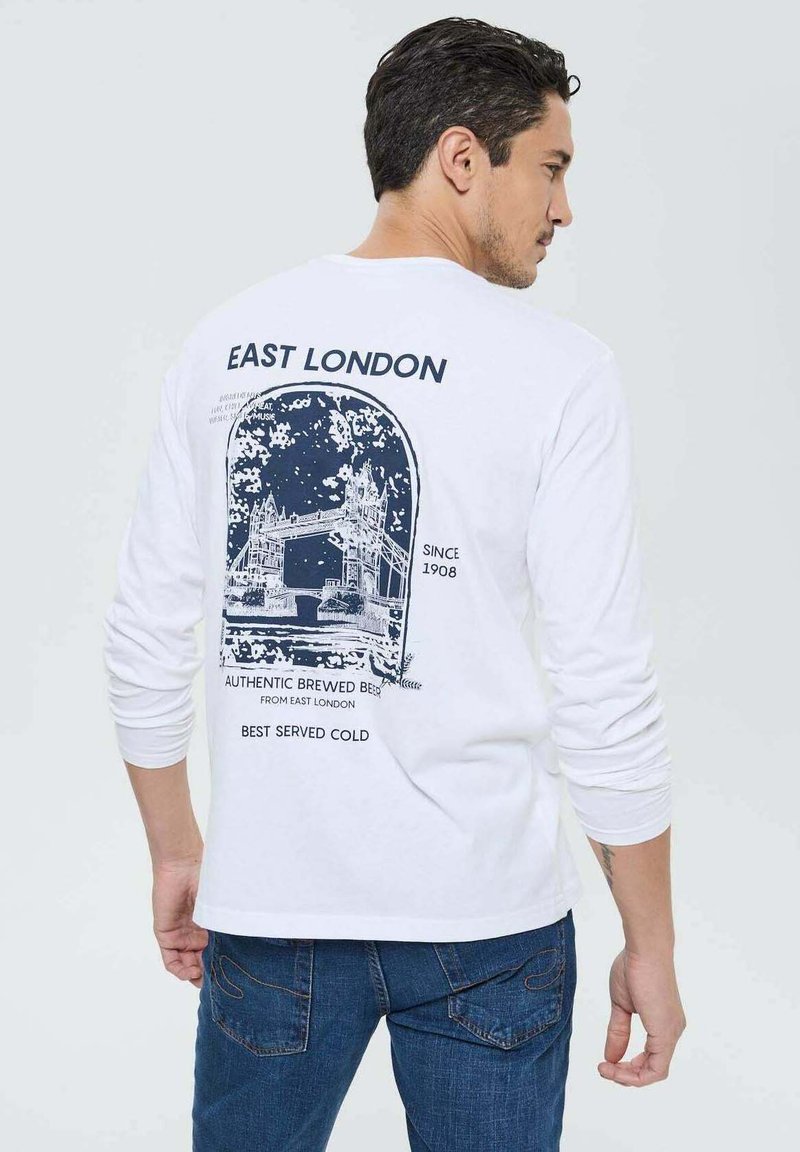 T-shirt à manches longues blanche avec un graphique navy du Tower Bridge et le texte "EAST LONDON" avec des détails de brasserie, porté avec un jean bleu.