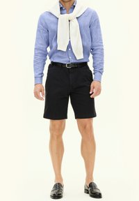 Jasnoniebieska w paski koszula na guziki, czarne szorty i czarne loafersy. Na szyi sweter w kolorze kremowym. Gładkie tkaniny i luźny krój.