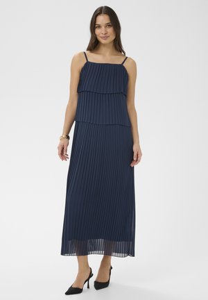 Femme portant une robe maxi plissée bleu marine à fines bretelles et des escarpins noirs, debout contre un fond clair uni.