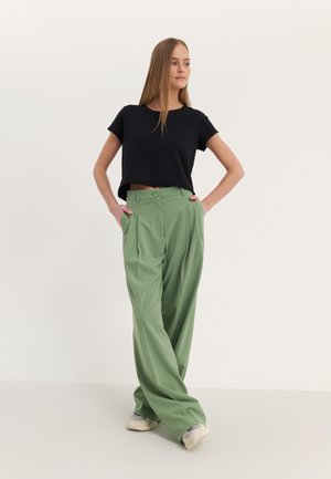 Vrouw draagt een zwart cropped t-shirt, brede groene broek en witte sneakers, staat met handen in de zakken, kijkt opzij.